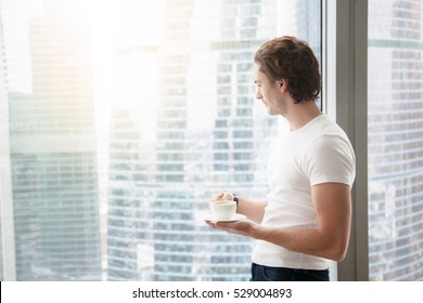 Young Handsome Carefree Man Near Modern Full Length Window Enjoying A Cup Of Coffee While Looking Outside, Good Morning, First Day In A New Office, Free From Work And Duty, Traveler. Copy Space