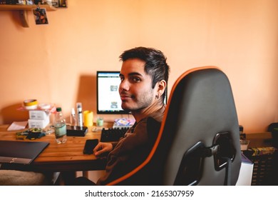 Young And Handsome Caucasian Boy With Earring In A Gamer Chair (in Gray And Orange Colors) In Front Of A Computer, Big Desk And Peach Wall