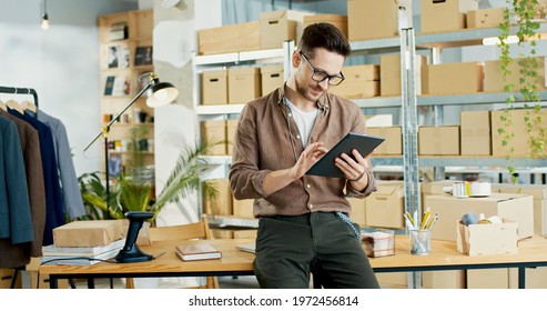 Young handsome Caucasian businessman in glasses with happy face typing on tablet at office in clothing shop. Man worker surfing internet on device in storehouse, business, delivery room - Powered by Shutterstock