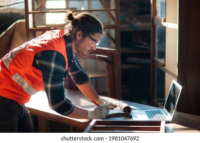 Young And Handsome Caucasian Carpenter In The Carpentry Shop Is Working And Checking Blueprint In Laptop To Repair A Furniture.Freelance Work At Home, Entrepreneur Or Small Business Owner. 