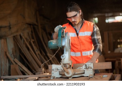 Young And Handsome Caucasian Carpenter Craftsman In Carpentry Shop Wearing Safety Uniform Workwear Use Circular Saws To Cut Wood Board To Make Furniture. Small Business And Freelance Craftsman