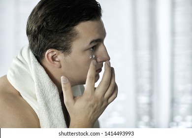Young handsome caucasian man applying cream under the eyes, towel on shoulders. Caring face, daily routine in the bathroom. Indoors, copy space. - Shutterstock ID 1414493993