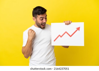 Young handsome caucasian man isolated on yellow background holding a sign with a growing statistics arrow symbol and celebrating a victory - Shutterstock ID 2164237051