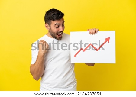 Young handsome caucasian man isolated on yellow background holding a sign with a growing statistics arrow symbol and celebrating a victory