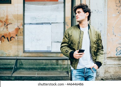 Young handsome caucasian man leaning on a bus stop holding a smart phone looking over and tapping the screen - technology, social network, communication concept