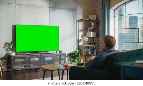 Young Handsome Caucasian Man Sitting On A Sofa And Watching TV With Horizontal Green Screen Mock Up. It's Day Time On Weekend At Home. Spacious Living Room In Stylish Loft Apartment.