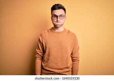 Young Handsome Caucasian Man Wearing Glasses And Casual Winter Sweater Over Yellow Background Puffing Cheeks With Funny Face. Mouth Inflated With Air, Crazy Expression.