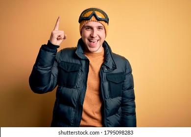 Young Handsome Caucasian Man Wearing Hat, Coat And Ski Glasses For Winter And Snow Weather Pointing Finger Up With Successful Idea. Exited And Happy. Number One.