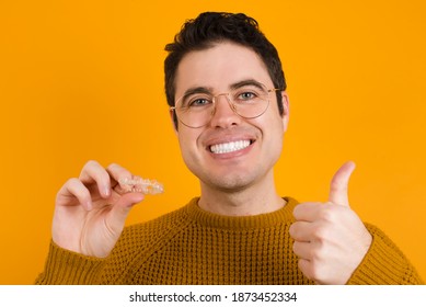 Young handsome Caucasian man wearing yellow sweater against orange wall holding a removable invisible aligner and rising thumb up making ok sign. 