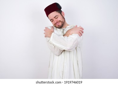 young handsome Caucasian man wearing Arab djellaba and Fez hat over white wall. Hugging oneself happy and positive, smiling confident. Self love and self care.