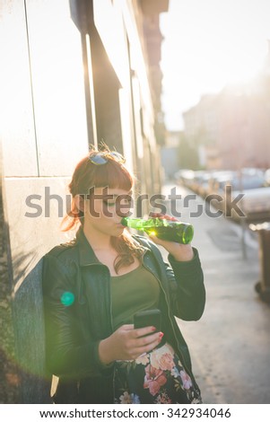Young handsome caucasian redhead woman drinking a beer and using a smartphone, looking down and tapping the screen - social network, technology, communication concept - backlight