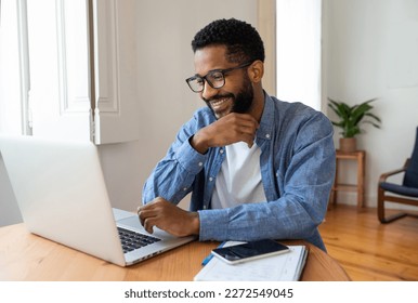 Young handsome cheerful black man working from home online remotely using his laptop. Looking at laptop screen, smiling.