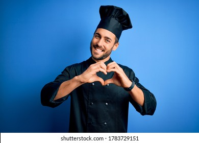 Joven y simpático chef, hombre con barba y uniforme de cocina y sombrero de fondo azul sonriendo enamorado con forma de símbolo cardíaco con las manos. Concepto romántico.