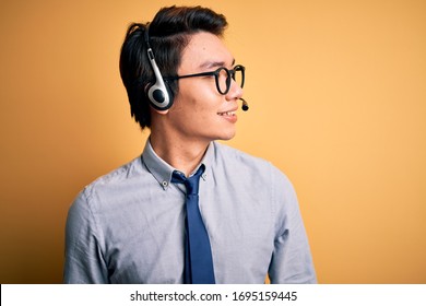 Young Handsome Chinese Call Center Agent Man Wering Glasses Using Headset Looking Away To Side With Smile On Face, Natural Expression. Laughing Confident.