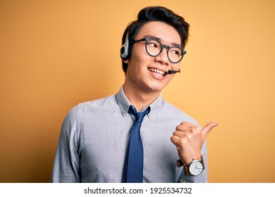 Young Handsome Chinese Call Center Agent Man Wering Glasses Using Headset Smiling With Happy Face Looking And Pointing To The Side With Thumb Up.