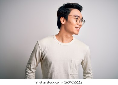 Young Handsome Chinese Man Wearing Casual T-shirt And Glasses Over White Background Looking Away To Side With Smile On Face, Natural Expression. Laughing Confident.