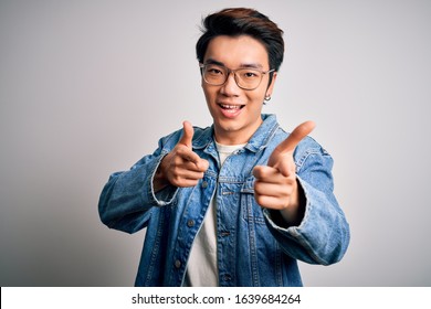 Young Handsome Chinese Man Wearing Denim Jacket And Glasses Over White Background Pointing Fingers To Camera With Happy And Funny Face. Good Energy And Vibes.