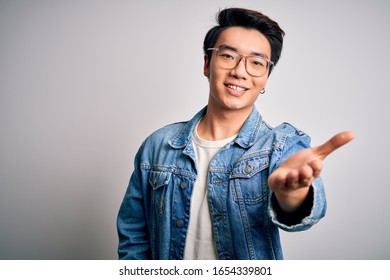 Young Handsome Chinese Man Wearing Denim Jacket And Glasses Over White Background Smiling Friendly Offering Handshake As Greeting And Welcoming. Successful Business.