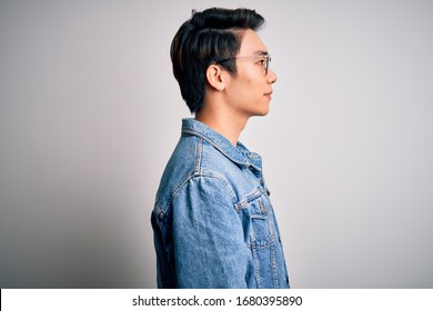 Young Handsome Chinese Man Wearing Denim Jacket And Glasses Over White Background Looking To Side, Relax Profile Pose With Natural Face With Confident Smile.