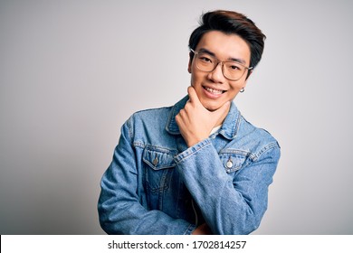 Young Handsome Chinese Man Wearing Denim Jacket And Glasses Over White Background Looking Confident At The Camera Smiling With Crossed Arms And Hand Raised On Chin. Thinking Positive.