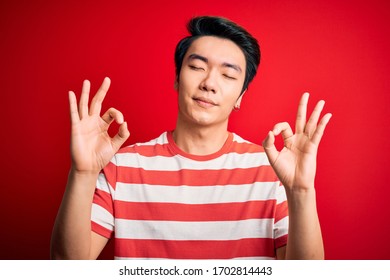 Young handsome chinese man wearing casual striped t-shirt standing over red background relaxed and smiling with eyes closed doing meditation gesture with fingers. Yoga concept. - Powered by Shutterstock