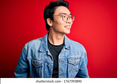 Young Handsome Chinese Man Wearing Denim Jacket And Glasses Over Red Background Looking Away To Side With Smile On Face, Natural Expression. Laughing Confident.