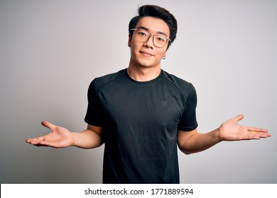 Young Handsome Chinese Man Wearing Black T-shirt And Glasses Over White Background Clueless And Confused Expression With Arms And Hands Raised. Doubt Concept.