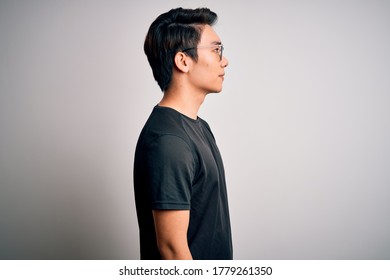 Young Handsome Chinese Man Wearing Black T-shirt And Glasses Over White Background Looking To Side, Relax Profile Pose With Natural Face With Confident Smile.