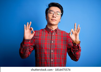 Young handsome chinese man wearing casual shirt and glasses over blue background relax and smiling with eyes closed doing meditation gesture with fingers. Yoga concept. - Powered by Shutterstock