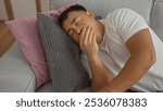 A young, handsome chinese man yawns while resting on a couch in his living room, showcasing a cozy indoor setting with pillows and comfortable home decor.