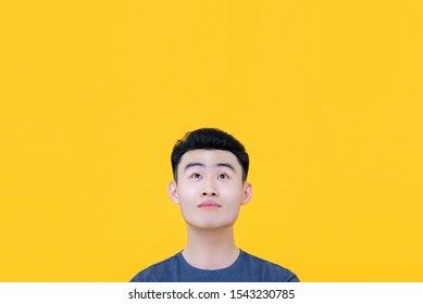 Young handsome clean shaven Asian man with thoughtful facial expression looking up to copy space with raised eyebrows isolated on yellow background