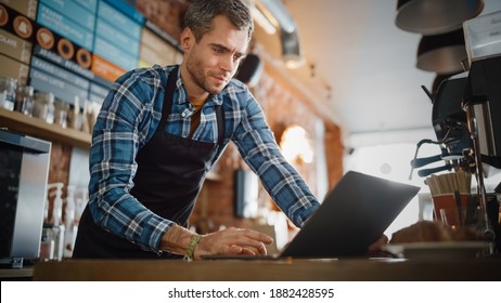 Young and Handsome Coffee Shop Owner is Working on Laptop Computer and Checking Inventory in a Cozy Cafe. Happy Restaurant Manager in Checkered Shirt Browsing Internet and Chatting with Friends.