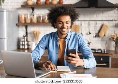 Young handsome confident African American casually dressed entrepreneur working from home using his mobile phone and laptop computer. Freelance, remote work, distant education concept. - Powered by Shutterstock