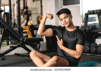 A young handsome and confident asian man flexing his bicep while sitting on the mat at the gym.