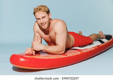 Young handsome cool sexy strong sporty blond man 20s wearing red shorts swimsuit lying on sup board paddle looking camera isolated on pastel blue background. Summer vacation sea rest sun tan concept.
