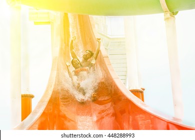 Young Handsome Couple Splashing Into A Waterslide In Aqua Park For Summer Vacation - Multiracial Friends Having Fun With Back Lighting - Holidays And Fun Concept - Soft Focus On Faces 