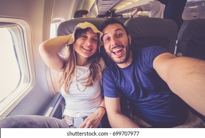 Young Handsome Couple Taking A Selfie On The Airplane During Flight Around The World. They Are A Man And A Woman, Smiling And Looking At Camera. Travel, Happiness And Lifestyle Concepts.