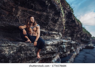 A young, handsome and courageous European man with drawings on his body sits on a background of rocks