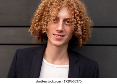 Young Handsome Curly Haired Ginger Male With Freckles Dressed In Stylish Casual Outfit Looking At Camera And Smiling Friendly Against Gray Wall