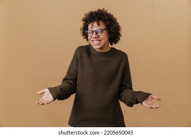 Joven y guapo chico alegre sonriente con gafas y suéter extendiendo las manos los lados y mirando la cámara  mientras está parado sobre aislado fondo beige