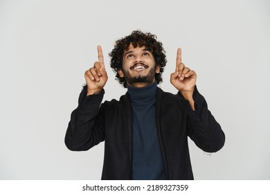 Young Handsome Curly Smiling Indian Man In Black Coat Pointing And Looking Upward Over Isolated Grey Background