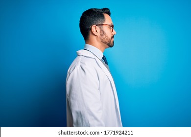 Young Handsome Doctor Man With Beard Wearing Coat And Glasses Over Blue Background Looking To Side, Relax Profile Pose With Natural Face With Confident Smile.