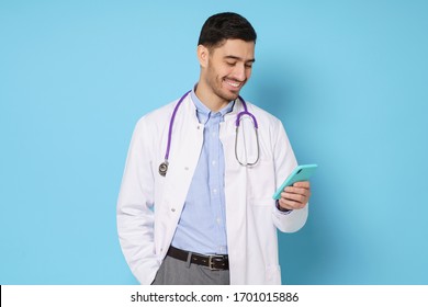 Young Handsome Doctor In White Coat Smiling While Looking At Screen Of His Phone, Using Medical App, Standing Isolated On Blue Background