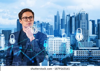 A young handsome eastern cybersecurity developer thinking about new concepts at security compliance division to protect clients confidential information. IT lock icons over Kuala Lumpur background.