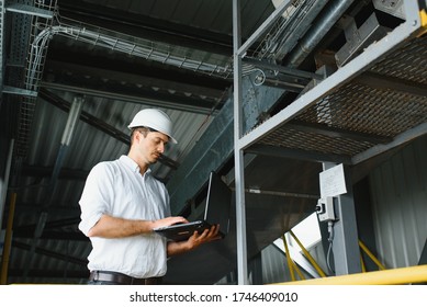 A Young Handsome Employee Of A Modern Factory In A White Hard. In The Background Is A Large Shop For The Production.