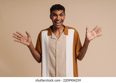 Young Handsome Enthusiastic African Man With Piercing With Raised Hands And Opened Mouth Looking At Camera While Standing Over Isolated Beige Background