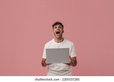 Young Handsome Enthusiastic Man With Opened Mouth Holding Laptop And Looking Upward , While Standing Over Pink Isolated Background