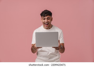 Young Handsome Enthusiastic Man With Opened Mouth Holding Laptop And Looking On It, While Standing Over Pink Isolated Background