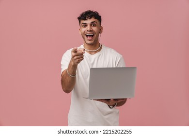 Young Handsome Enthusiastic Man With Opened Mouth Holding Laptop, Pointing And Looking At Camera, While Standing Over Isolated Pink Background