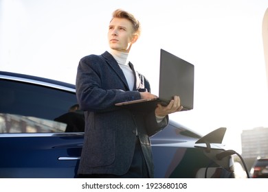 Young Handsome European Businessman At Charging Station, Standing Near Electric Car With Laptop Pc And Looking Away, While Refueling His Auto. EV Station And People Concept